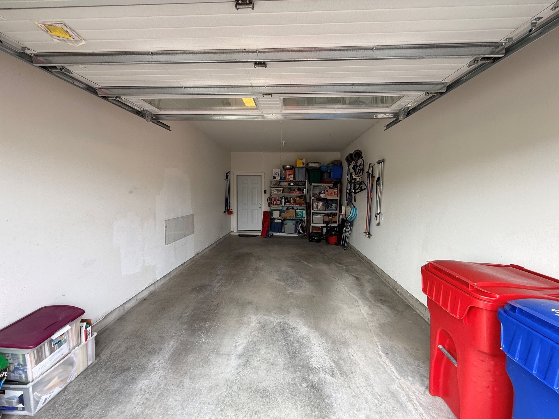 Empty garage interior with concrete floor, white walls, and red and blue storage bins on the right.