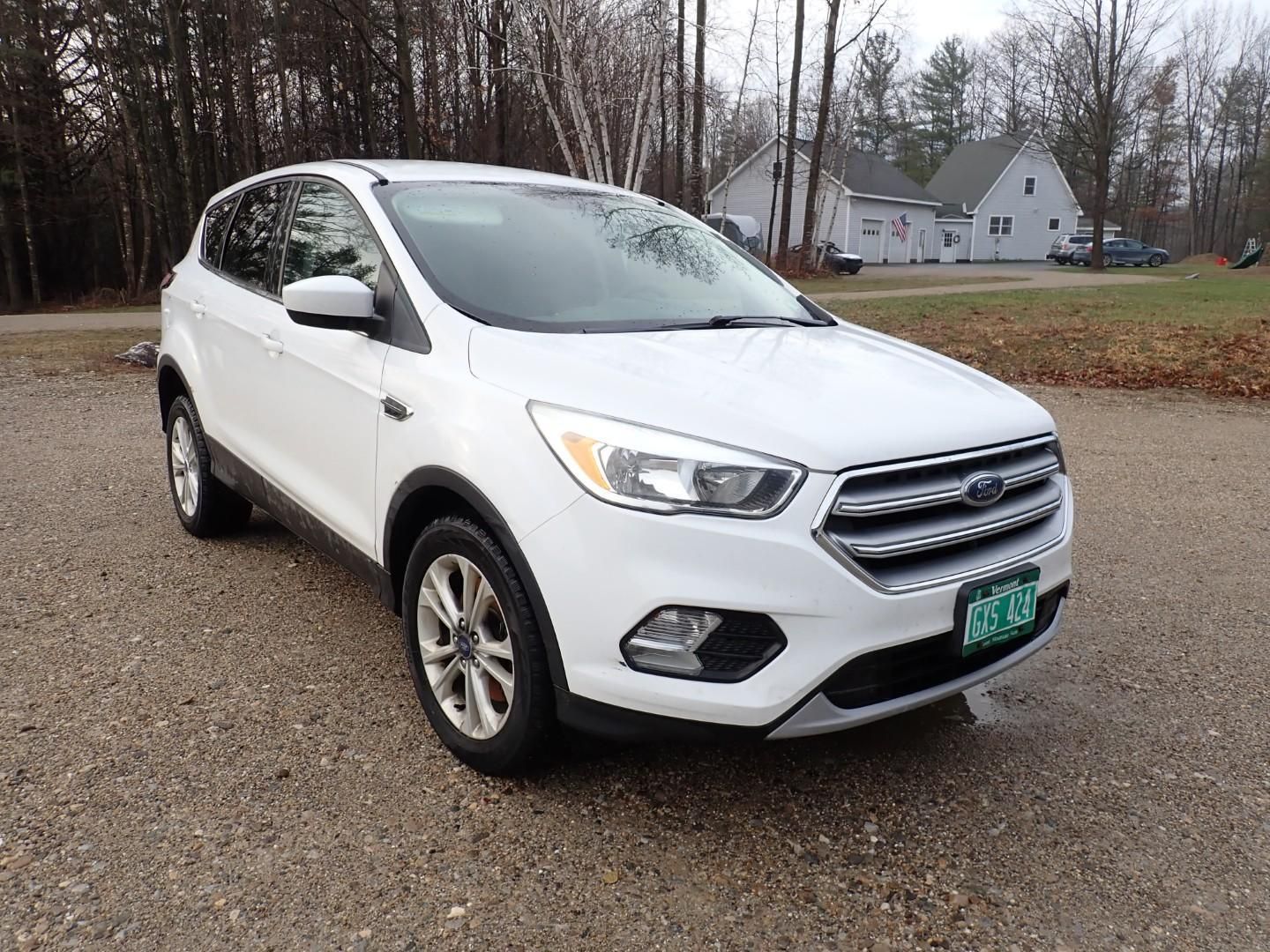 A white Ford Escape SUV parked on a gravel surface outdoors with trees and a house in the background.
