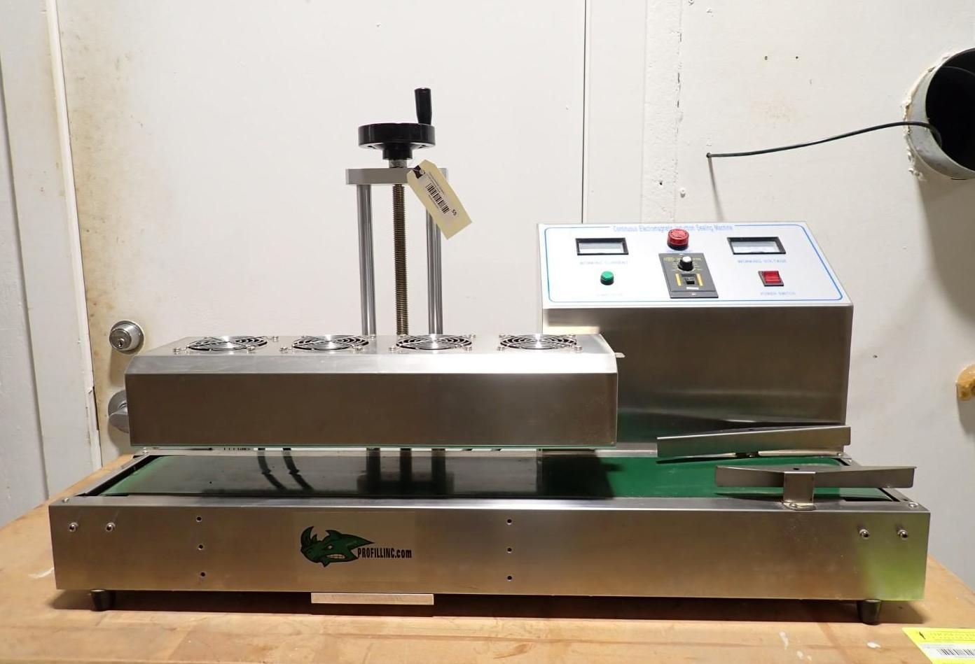 A stainless steel industrial conveyor belt induction sealing machine with a control panel, sitting on a wooden surface.