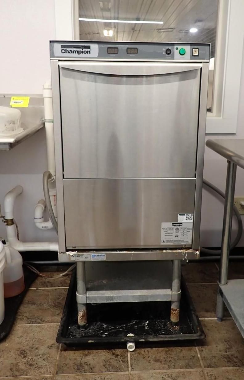 A commercial stainless steel under-counter dishwasher sits on a black drain pan in a tiled kitchen area.