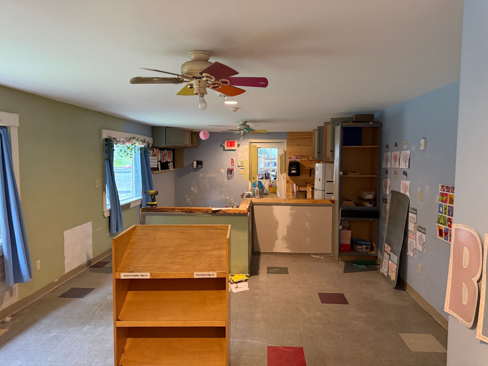 Empty classroom with shelves, a front counter, blue walls, and a ceiling fan on a tiled floor.