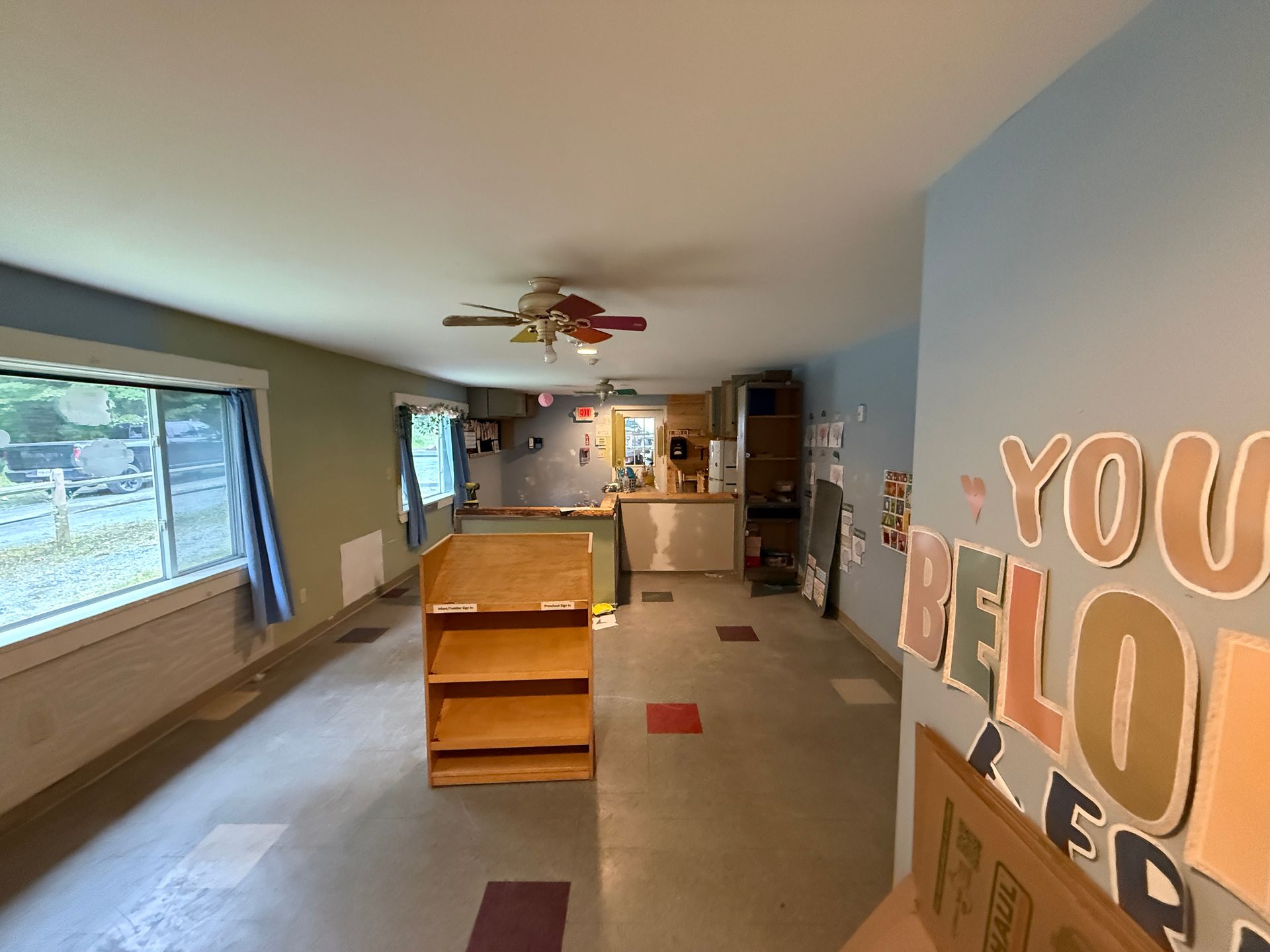 Wide living room with window seat, ceiling fan, and blue wall with large “YOU BELONG HERE” letters