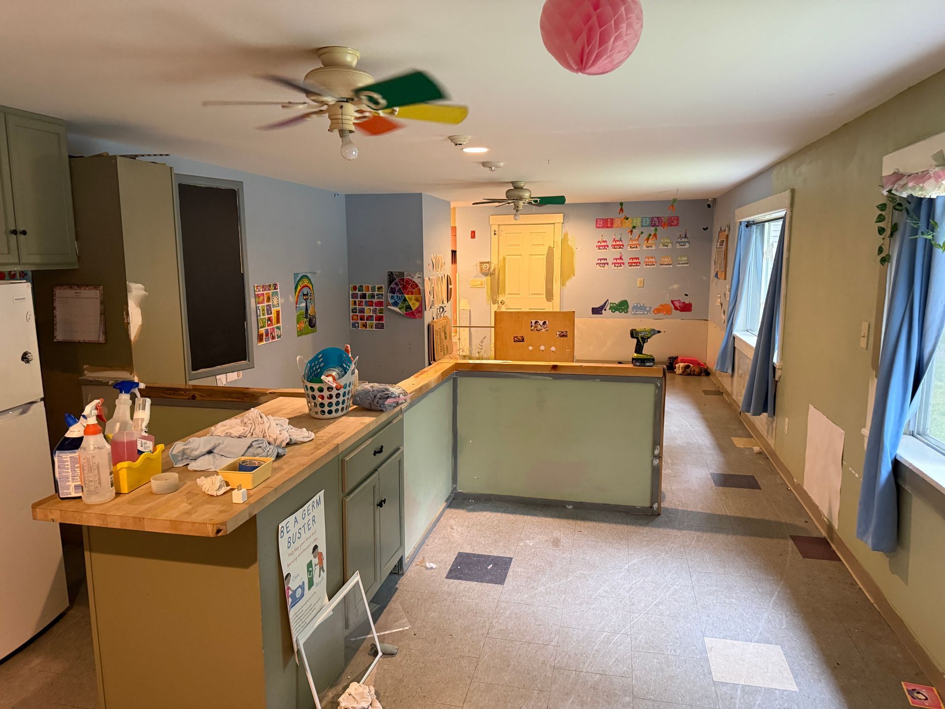 Bright classroom with counters, art supplies, wall decorations, and a ceiling fan; empty and well lit.