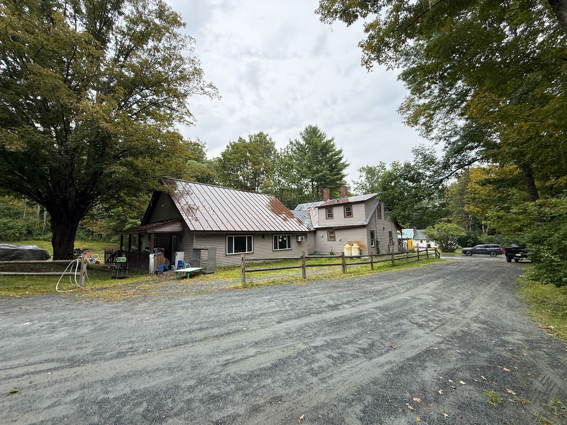 Rural gravel road leading past a rustic house and trees under a cloudy sky
