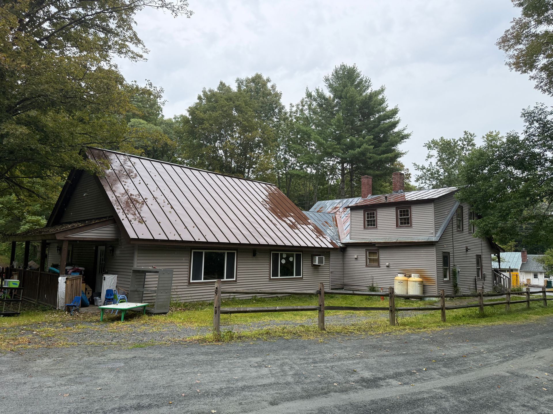 Rustic gray cabin and barn with metal roofs beside a gravel road, surrounded by trees and a fence