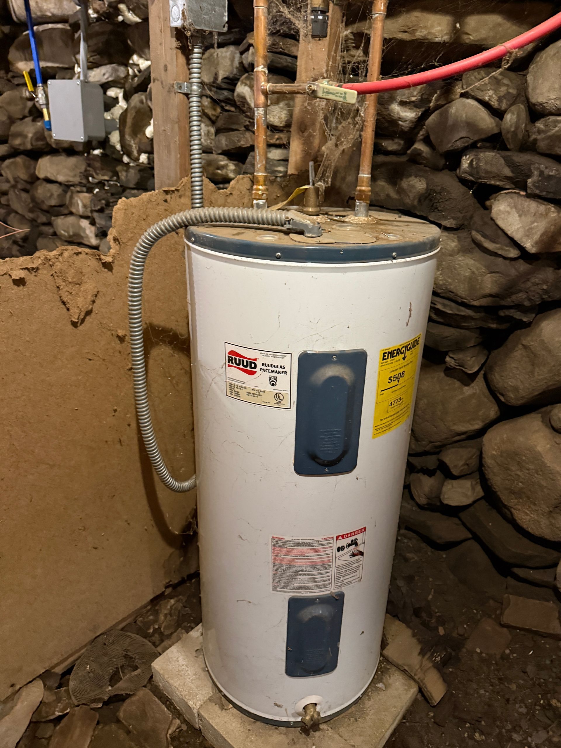 White water heater tank with pipes and valves in a stone-walled utility area.