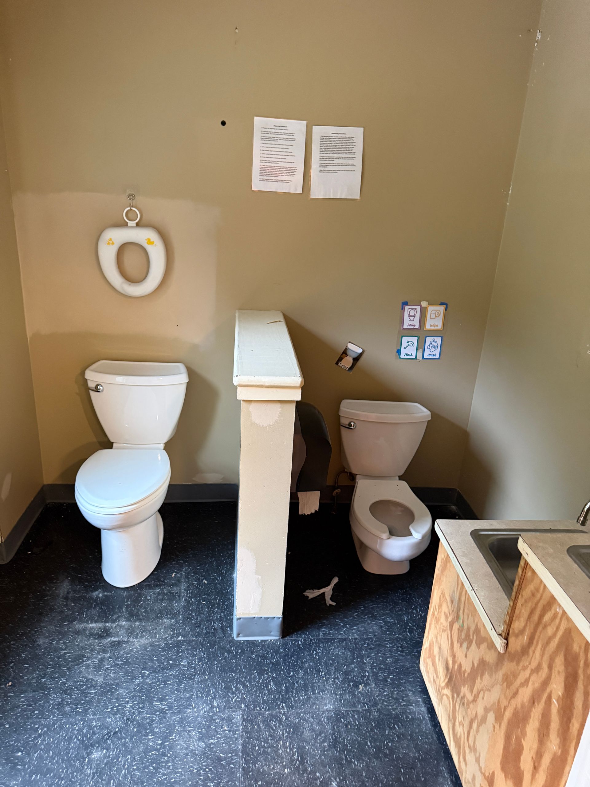 restroom with two toilets, a wall-mounted toilet paper holder, and beige walls with posted signs.
