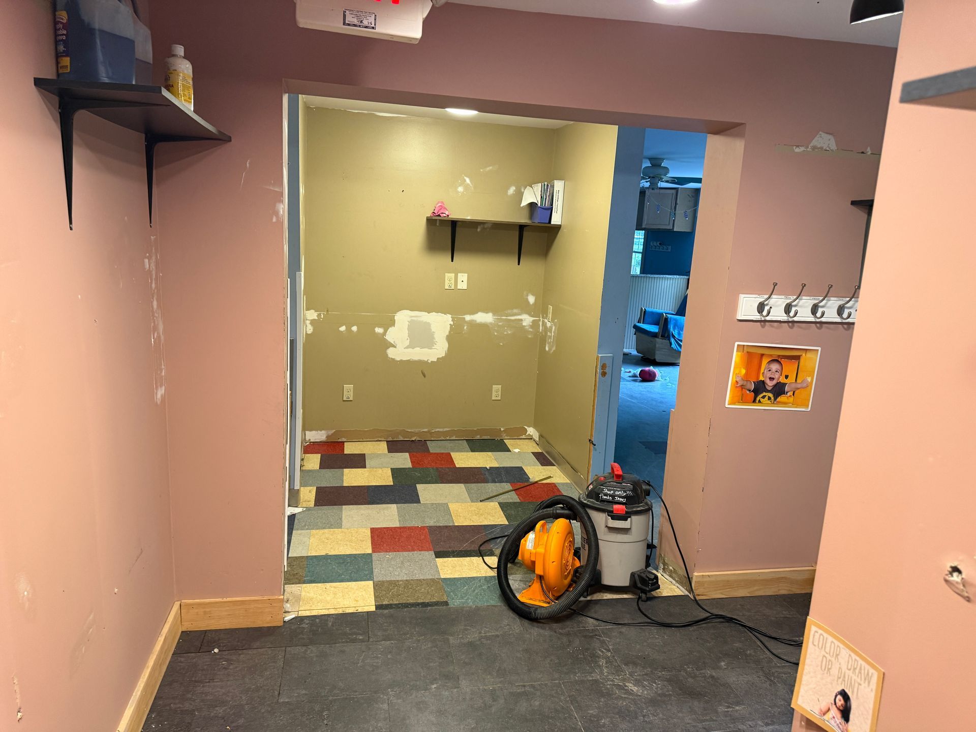 Vacant room with pink walls, doorway to a tile-floored back room, and a vacuum on the carpet.