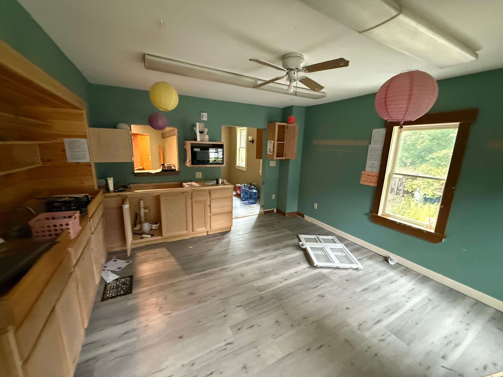 Empty teal living room with kitchen counter, wood floors, ceiling fan, and hanging paper lanterns
