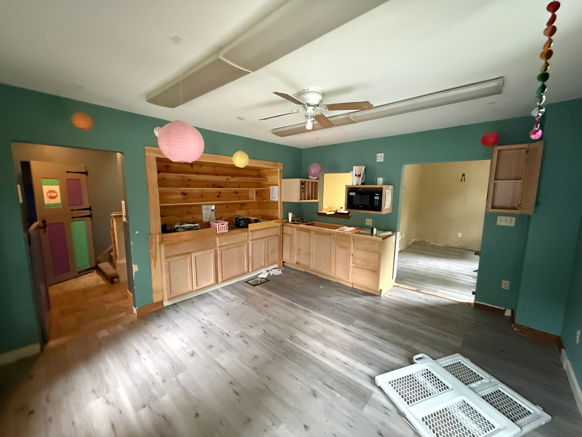 Empty kitchen with teal walls, wood cabinets, and sunlight on gray floorboards.