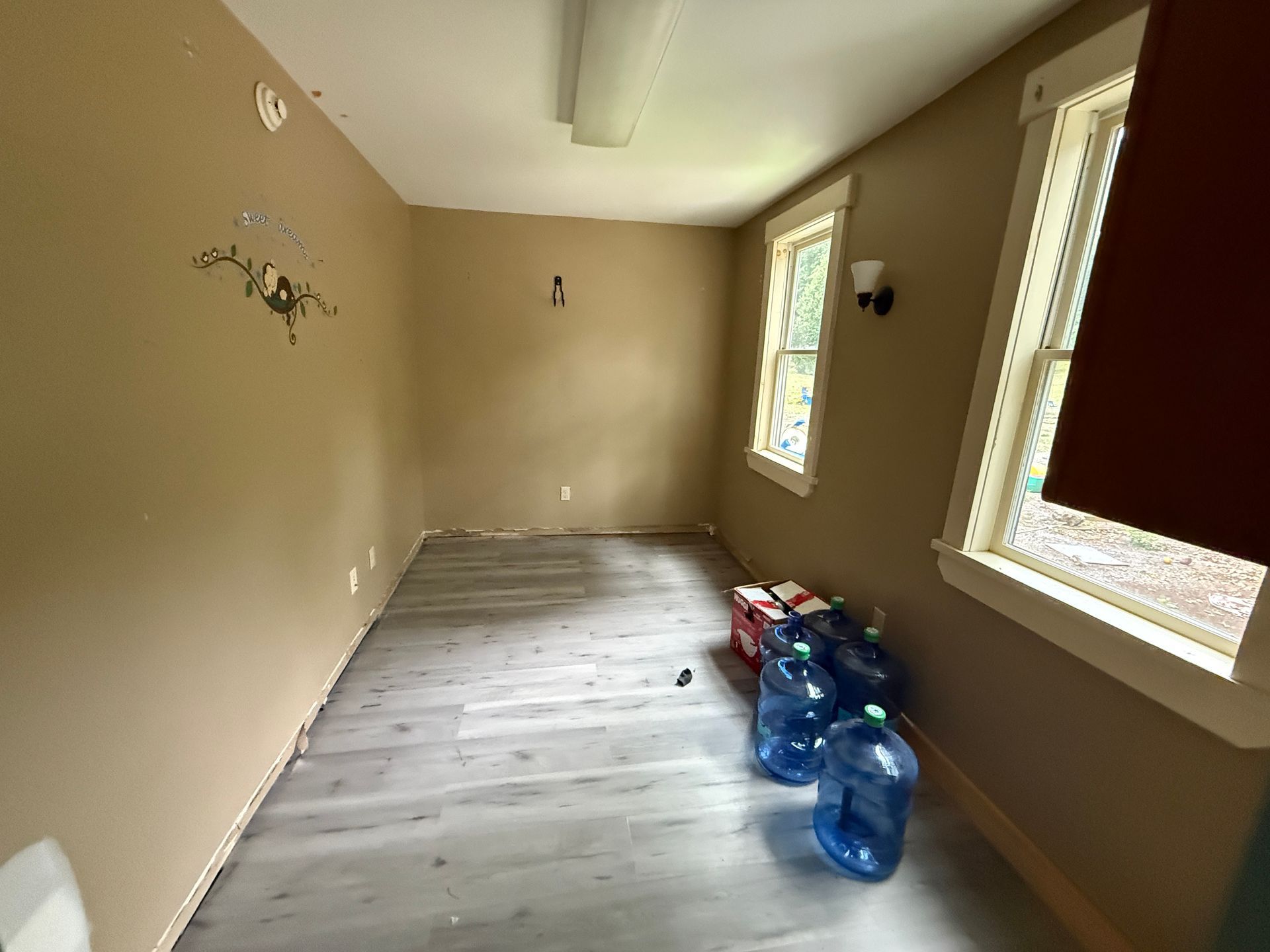 Empty beige room with light wood floor, windows on the right, and water jugs stacked near the far wall