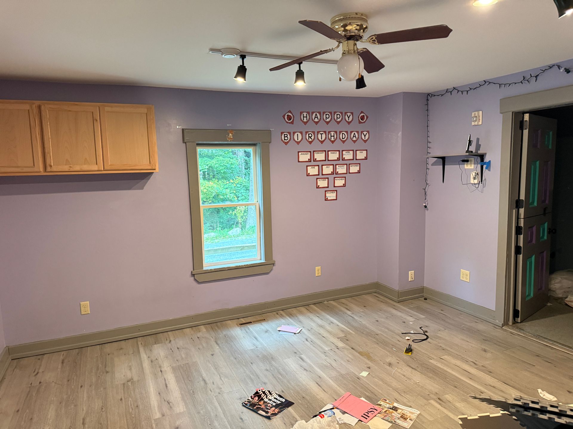 Empty purple room with wood floor, ceiling fan, wall cabinets, and a single window.