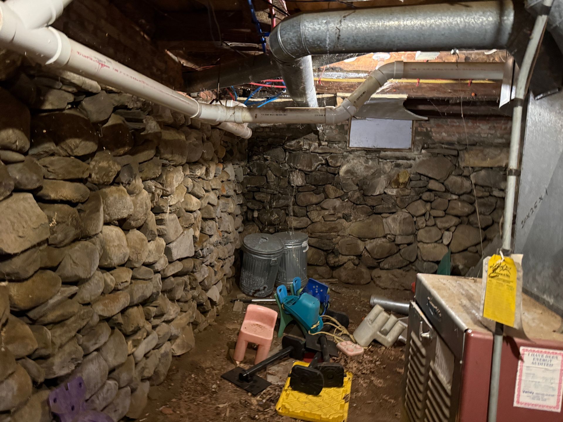 Basement crawlspace with stone walls, exposed pipes, and cleaning tools