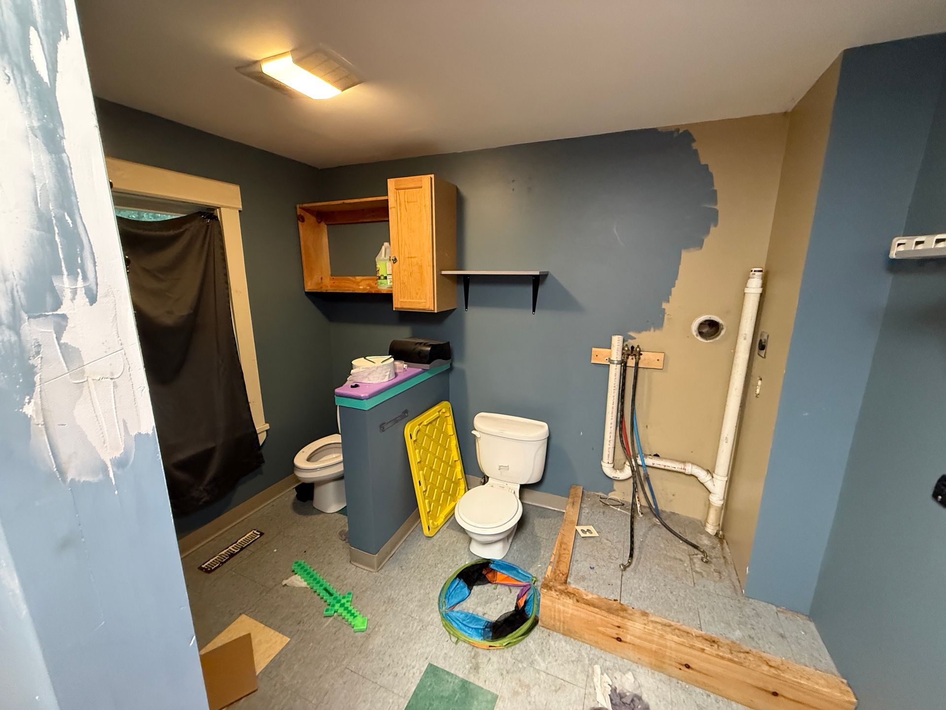 Small bathroom under renovation with blue walls, exposed plumbing, toilet, sink cabinet, and scattered tools.