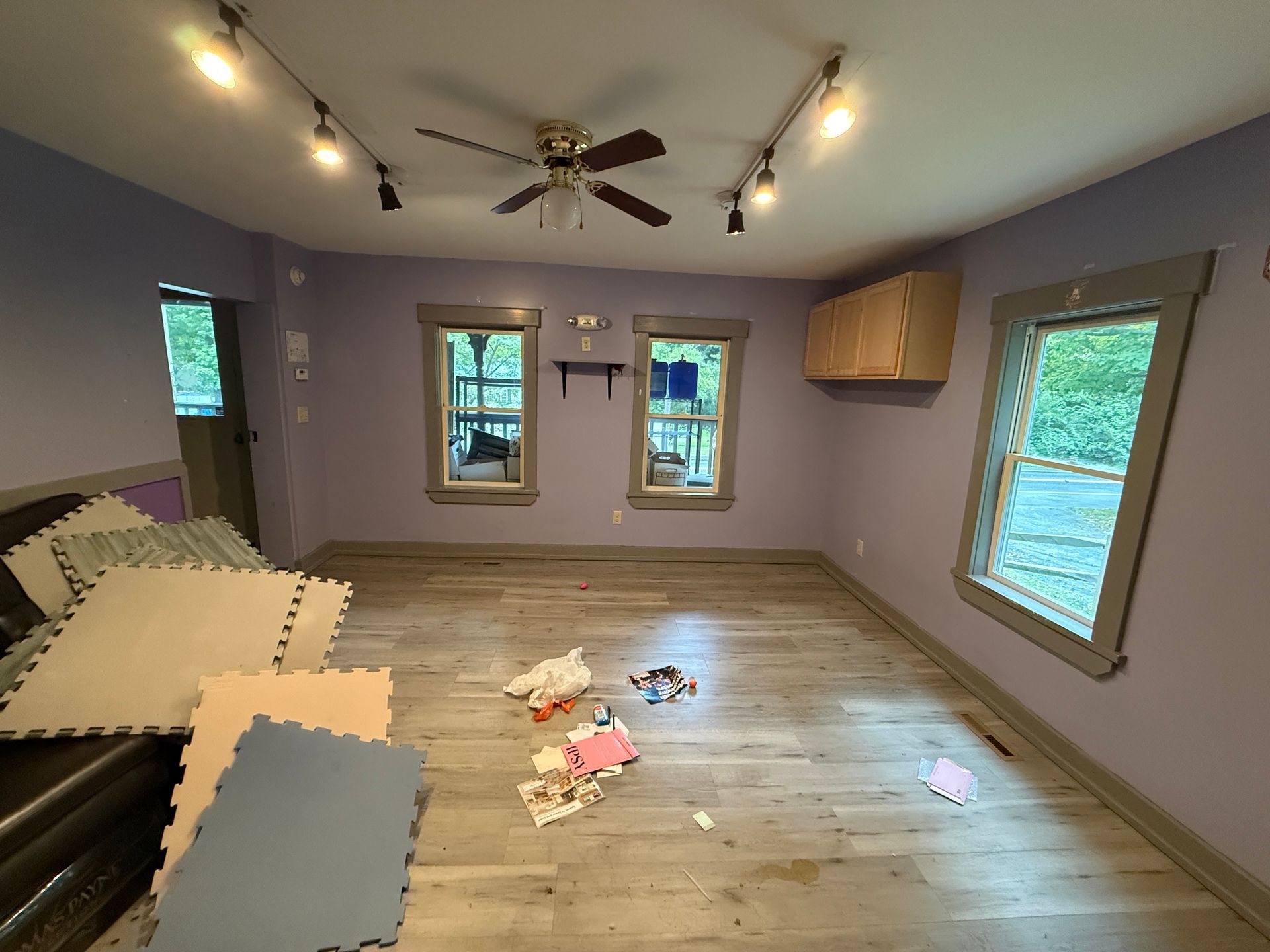 Empty playroom with light purple walls, wood floor, windows, foam mats, and toys scattered on the floor