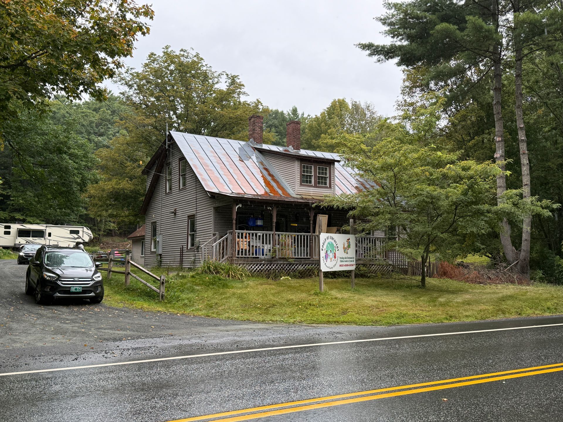 Rustic cabin by a road with a parked car, surrounded by trees and greenery