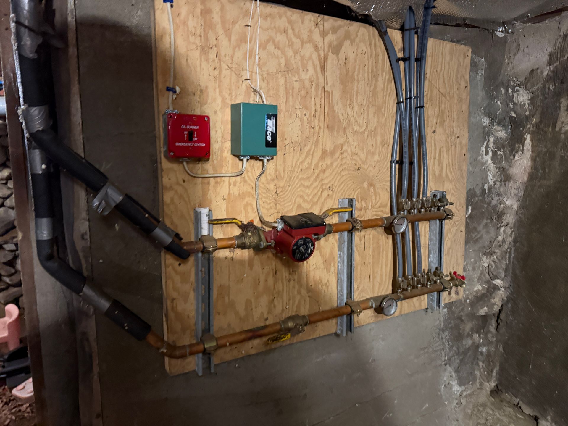 Basement utility wall with pipes, valves, and red and green electrical boxes mounted on plywood backing.