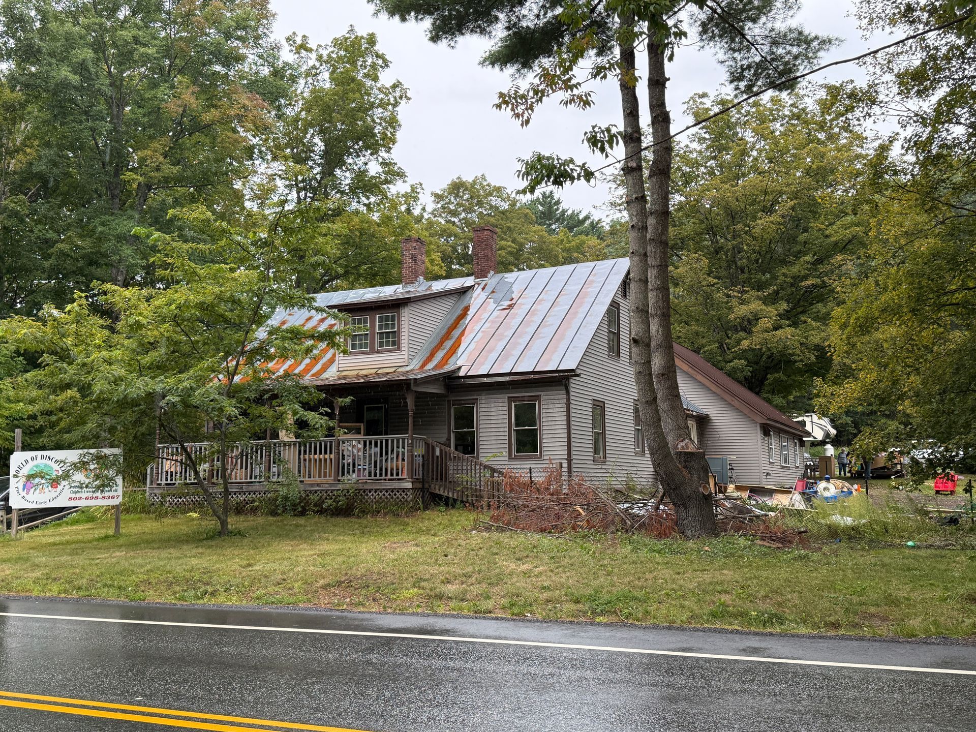 rustic house with a metal roof beside a wet road, surrounded by trees and grass