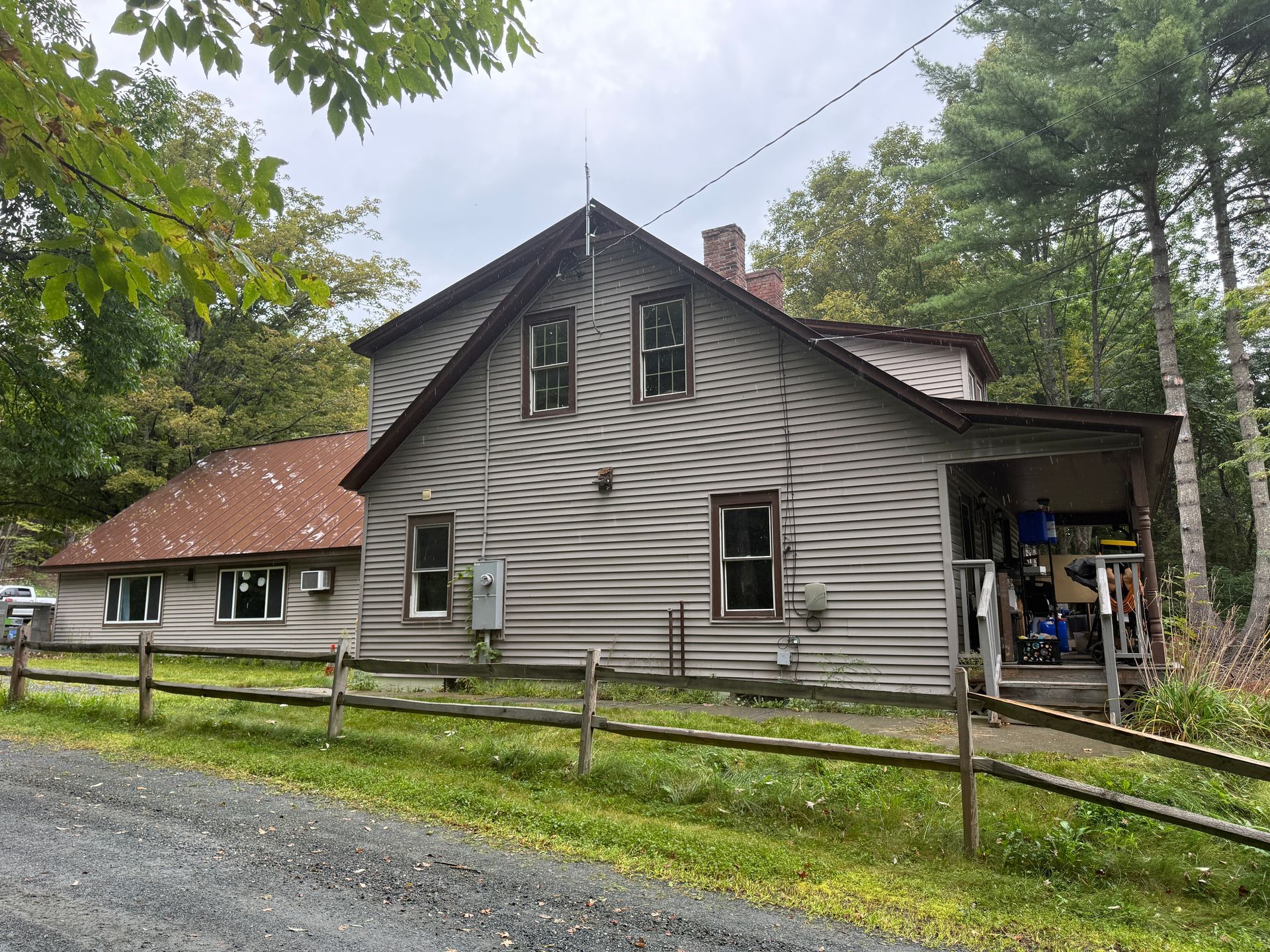 gray farmhouse with red roof beside a gravel drive, surrounded by trees and a wooden fence.