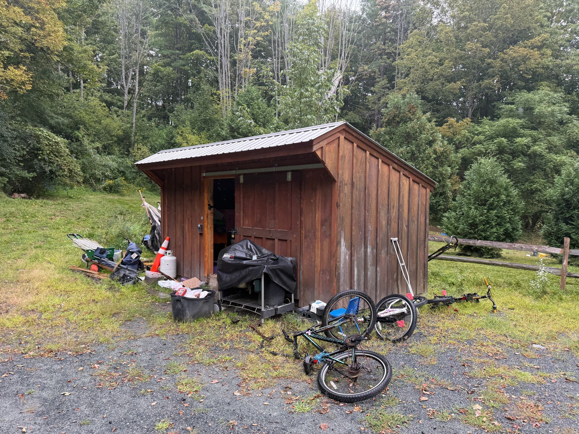 Small brown shed in a wooded yard, with bicycles and assorted items