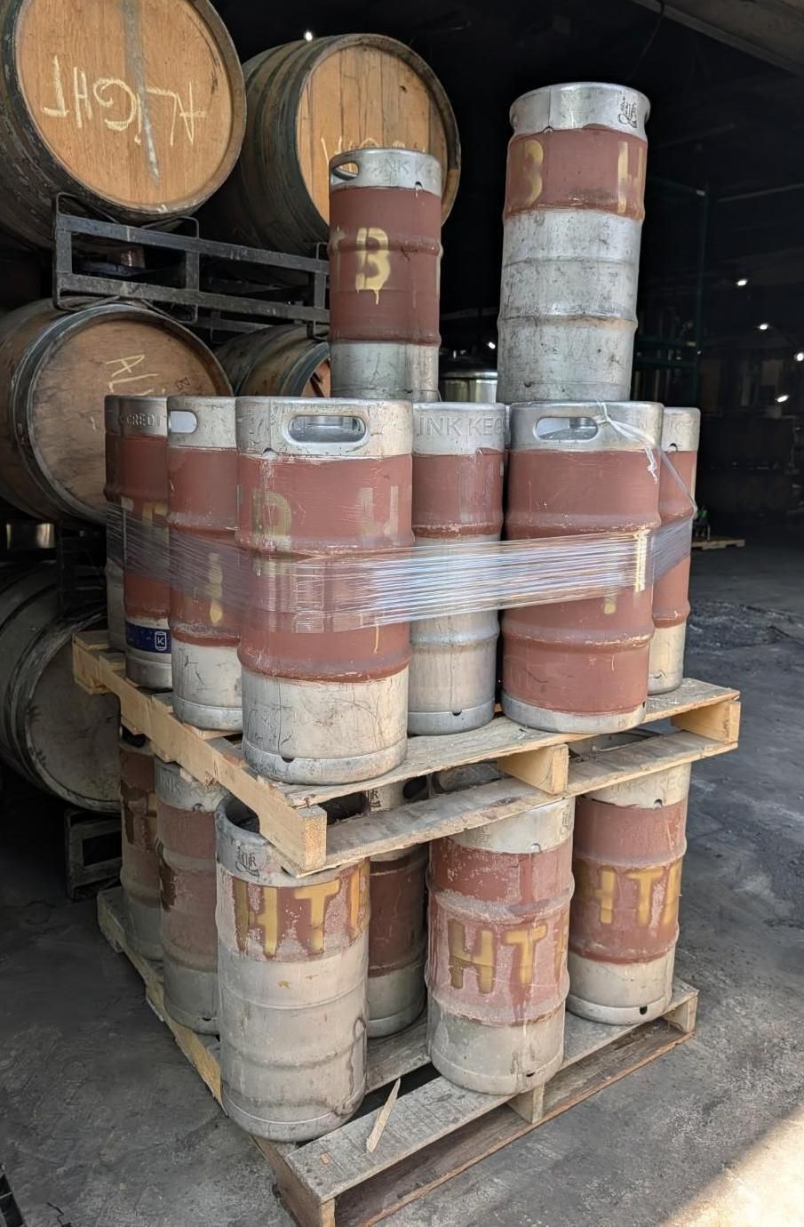 Stacked metal drums and barrels on wooden pallets inside a dark warehouse.