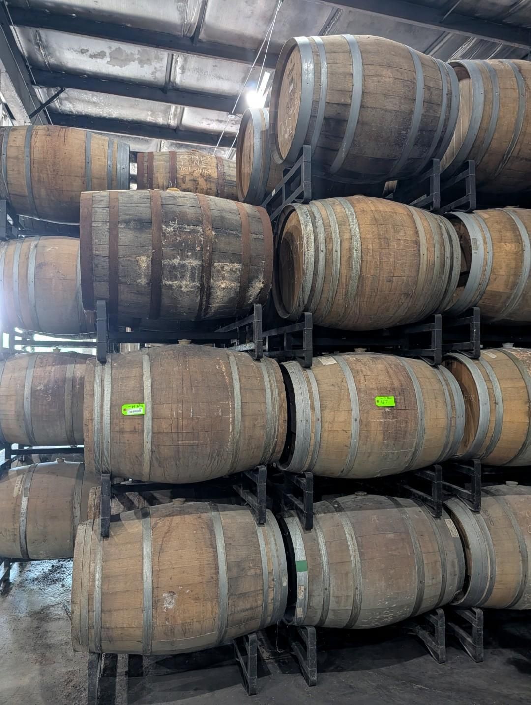 Stacked wooden wine barrels in a storage warehouse under a metal roof.