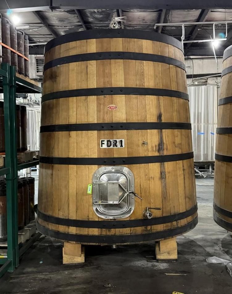 Large wooden industrial tank with black bands and a metal hatch in a warehouse.