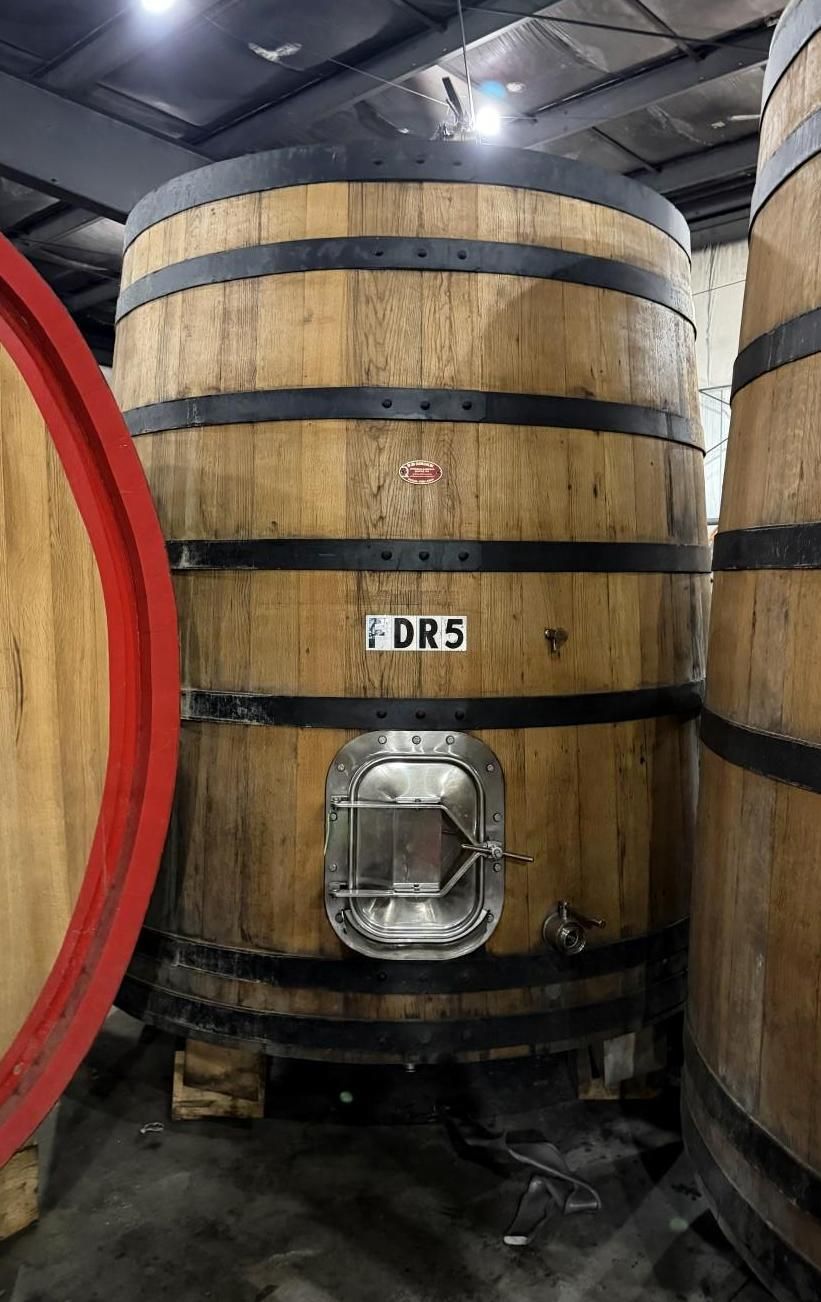 Large wooden wine barrel in a cellar, with metal bands and a small hatch labeled “DRS”.