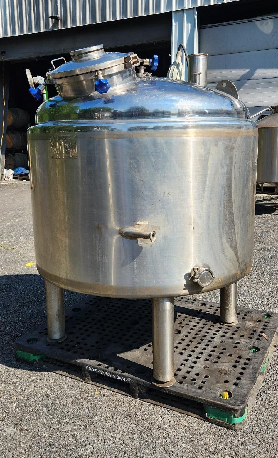 Stainless steel industrial tank on legs outdoors, with a top hatch and front spout.