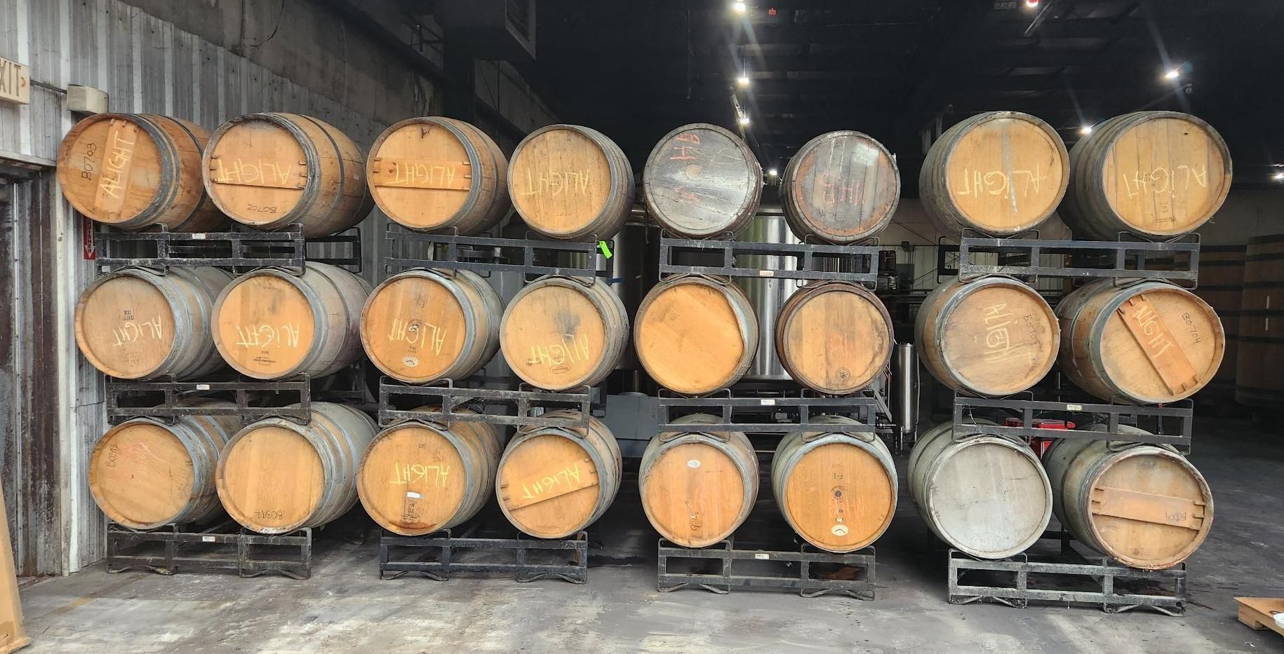 Rows of wooden wine barrels stacked in a cellar, with dark metal racks and a concrete floor.