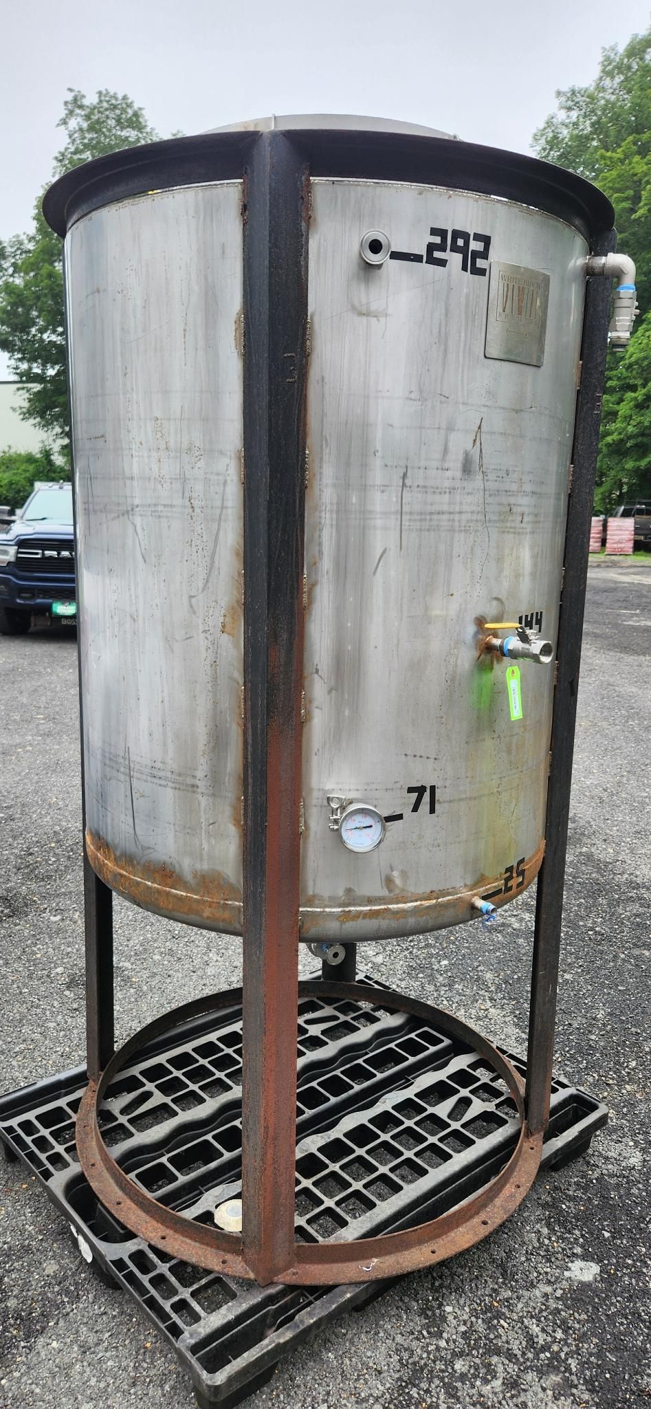 Large stainless steel tank on a metal stand, outdoors on gravel near trees.
