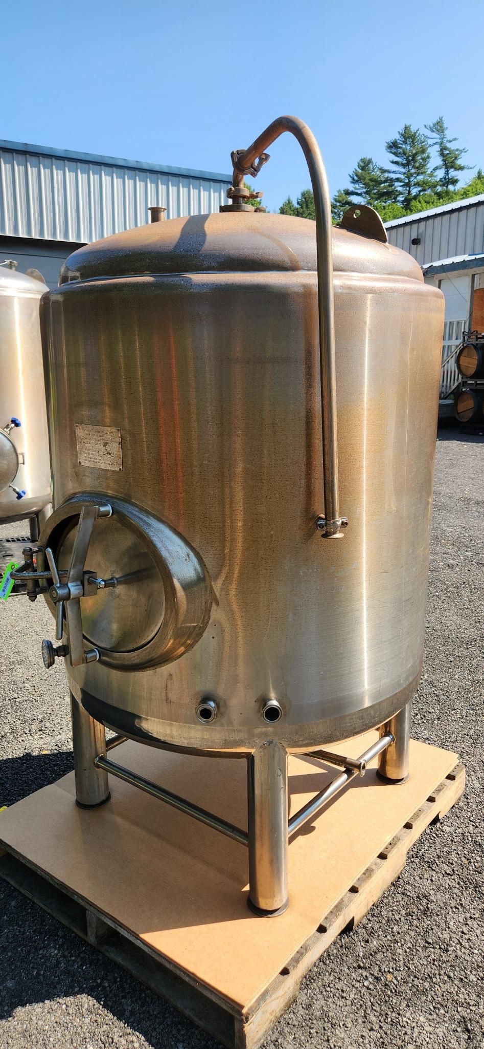 Stainless steel tank on a wooden pallet outdoors, with a side valve and top pipe handle.