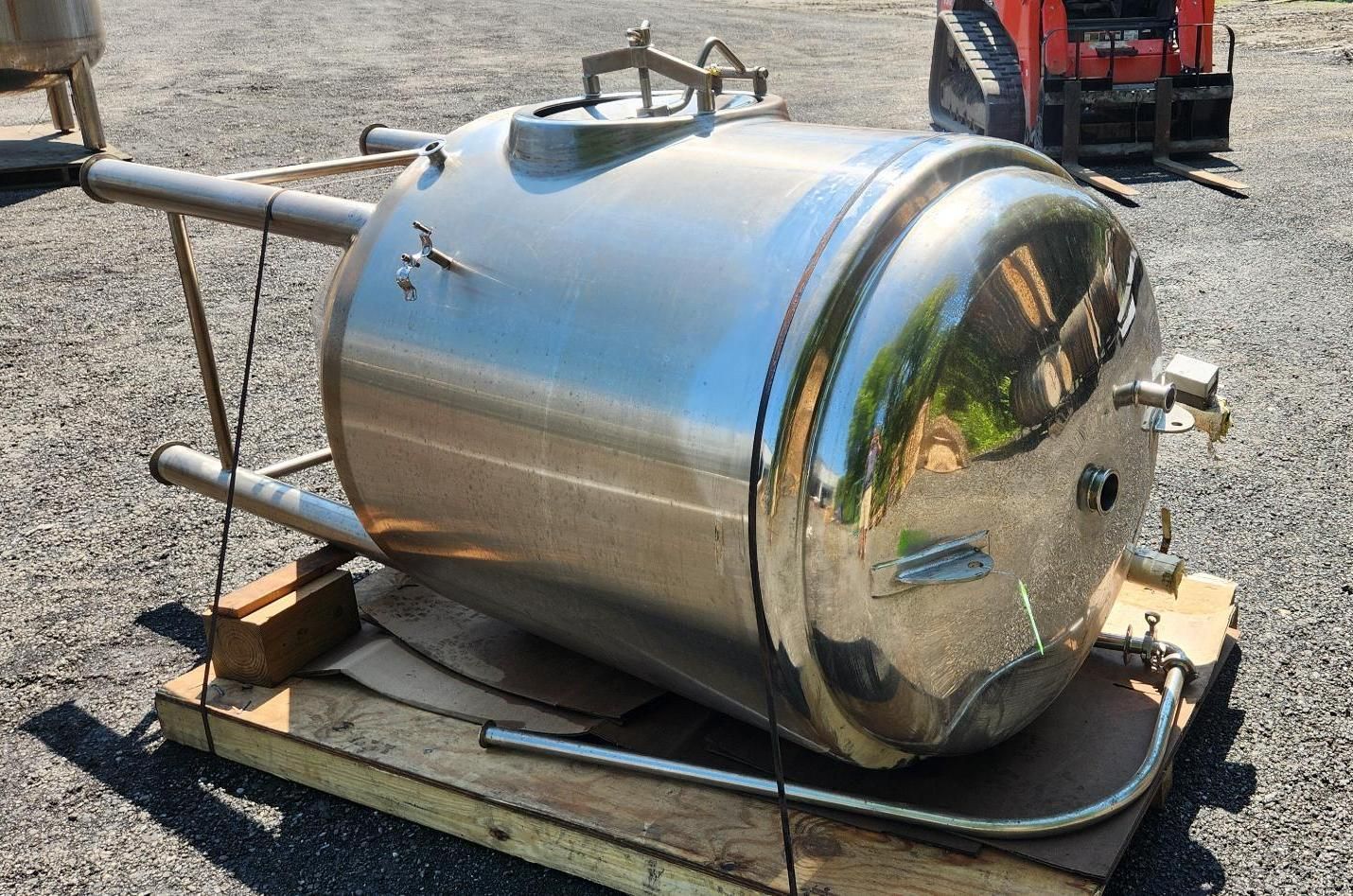 Stainless steel tank on a wooden pallet in a gravel lot, with pipes and a handle on top.