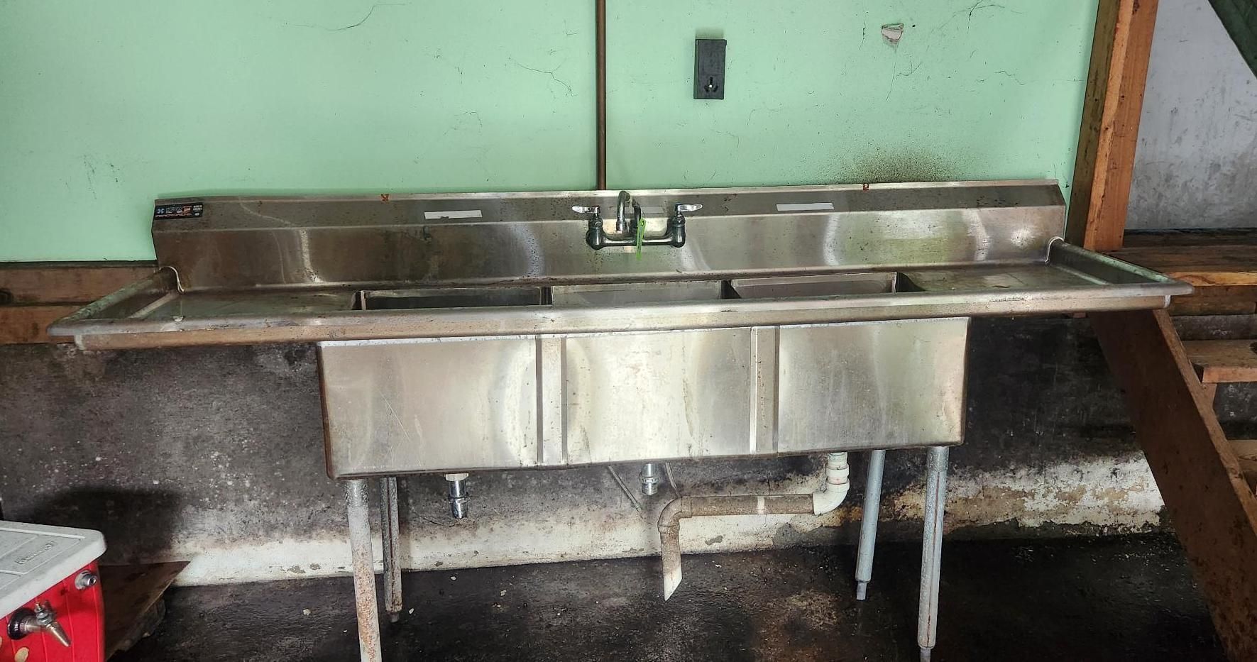 Stainless steel triple-sink counter in a kitchen or utility room, with drain pipes visible underneath