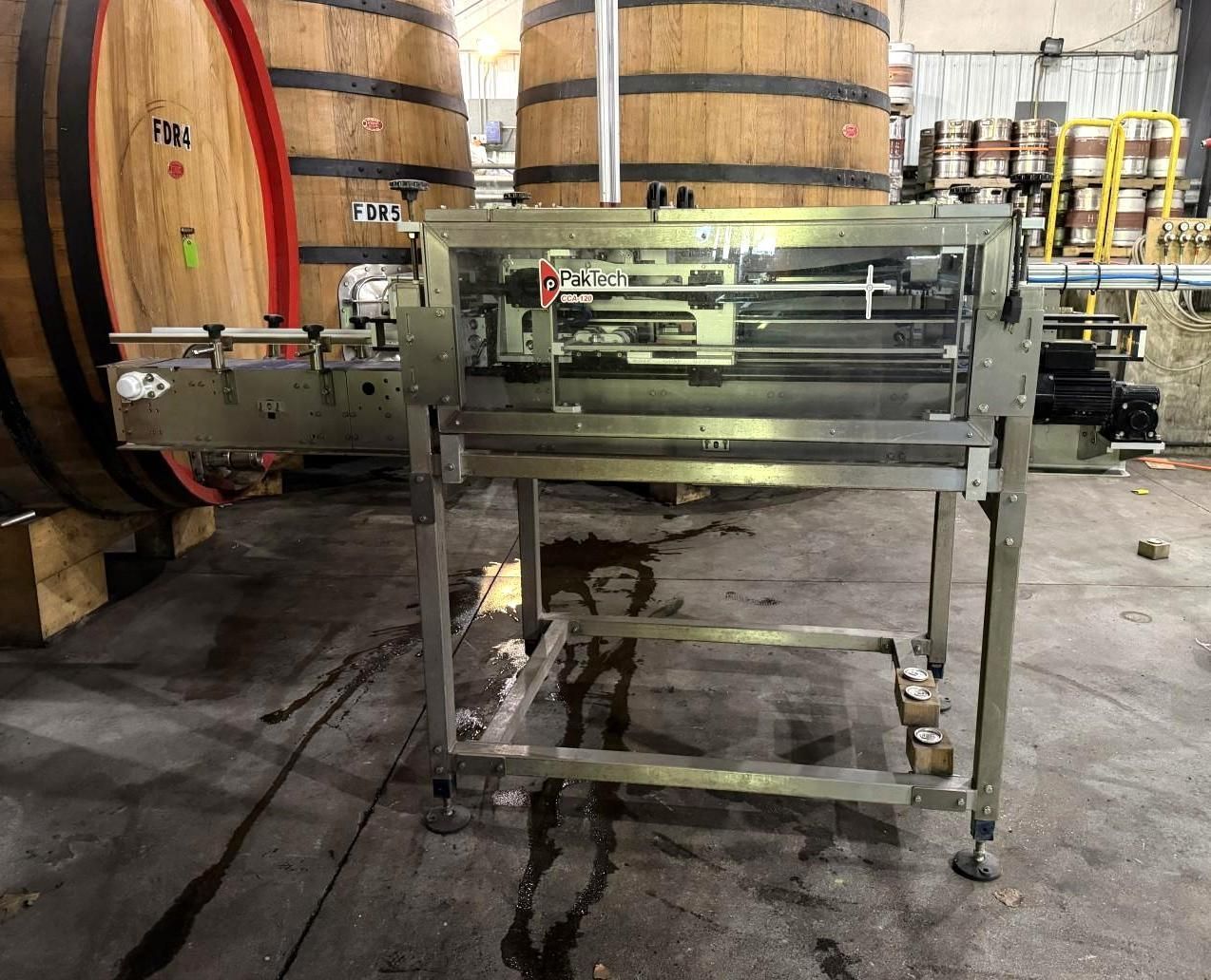 Stainless steel bottling machine in a winery, with wooden barrels in the background.