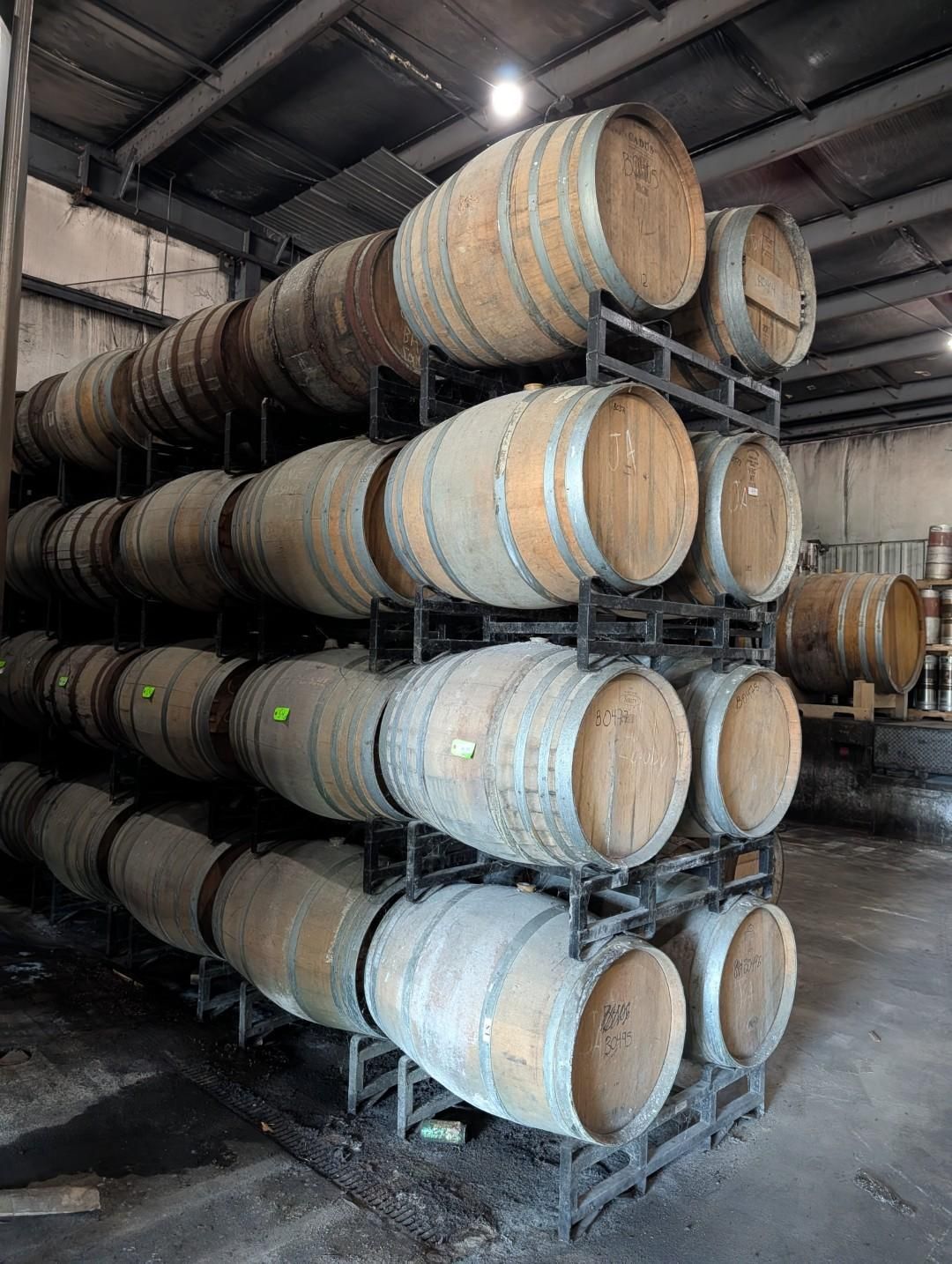Stacked wooden barrels in an industrial storage room, with metal racks and concrete floor.