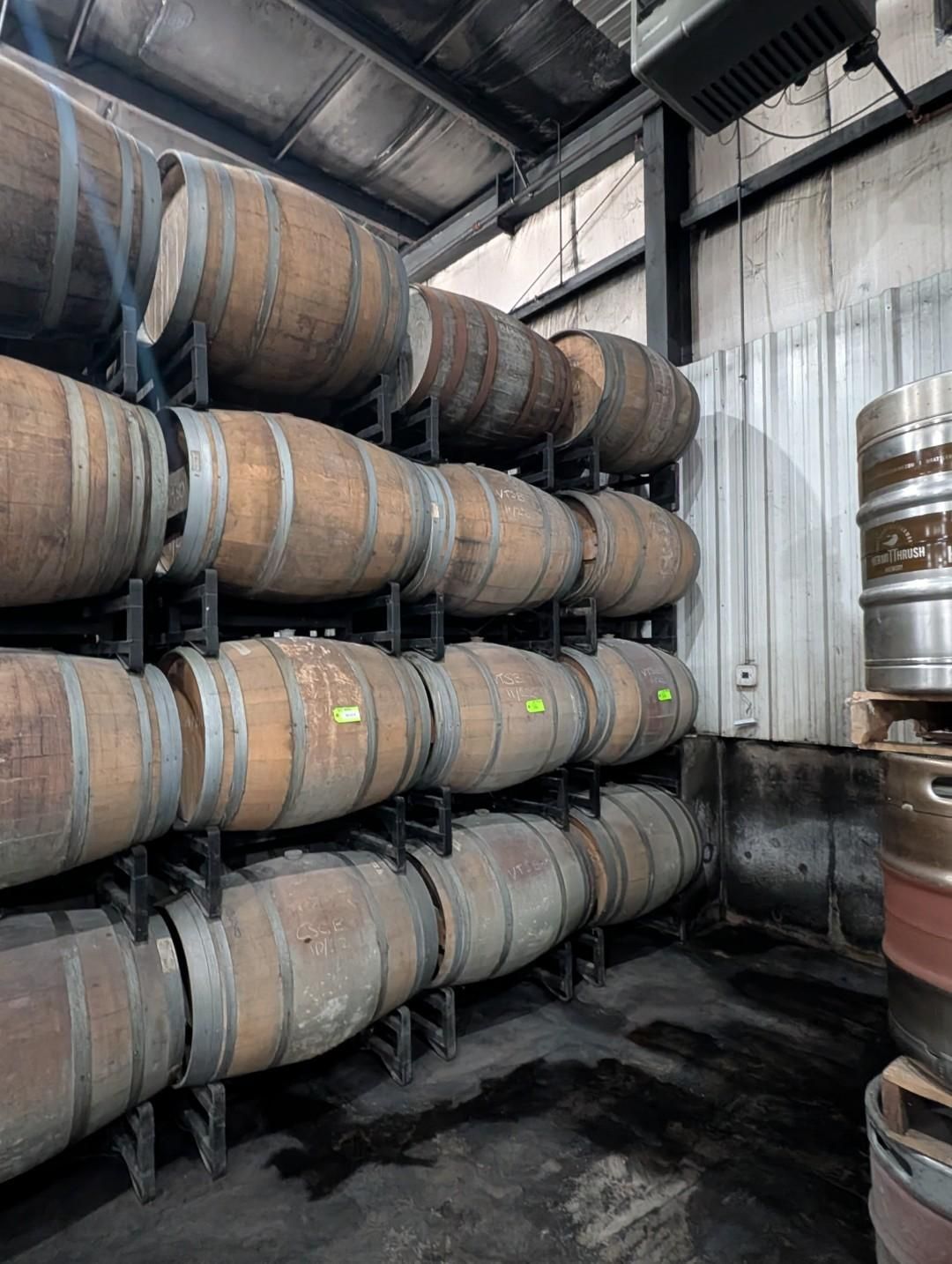 Rows of wooden barrels stacked in a warehouse cellar, with one metal keg on the right.