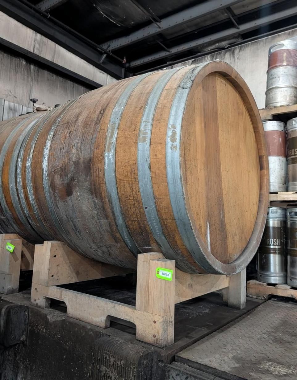 Large wooden wine barrel on a stand in an industrial warehouse, with stacked barrels in the background.