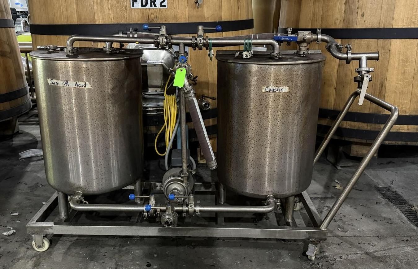 Two large stainless steel brewing tanks on a wheeled metal frame in an industrial setting.