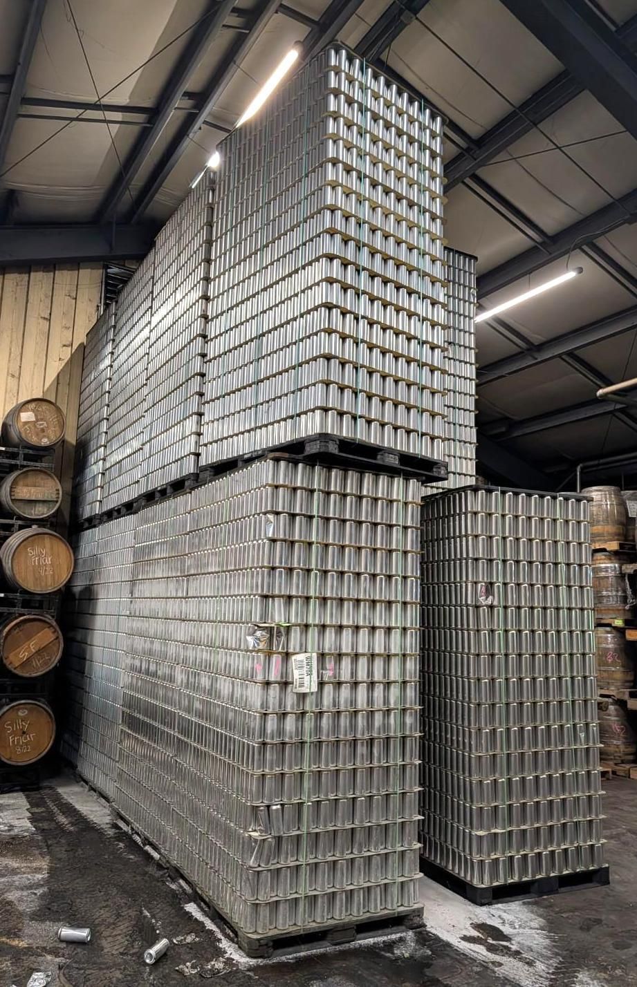 Stacked silver metal cans in a warehouse, arranged in tall pallets under an industrial ceiling.