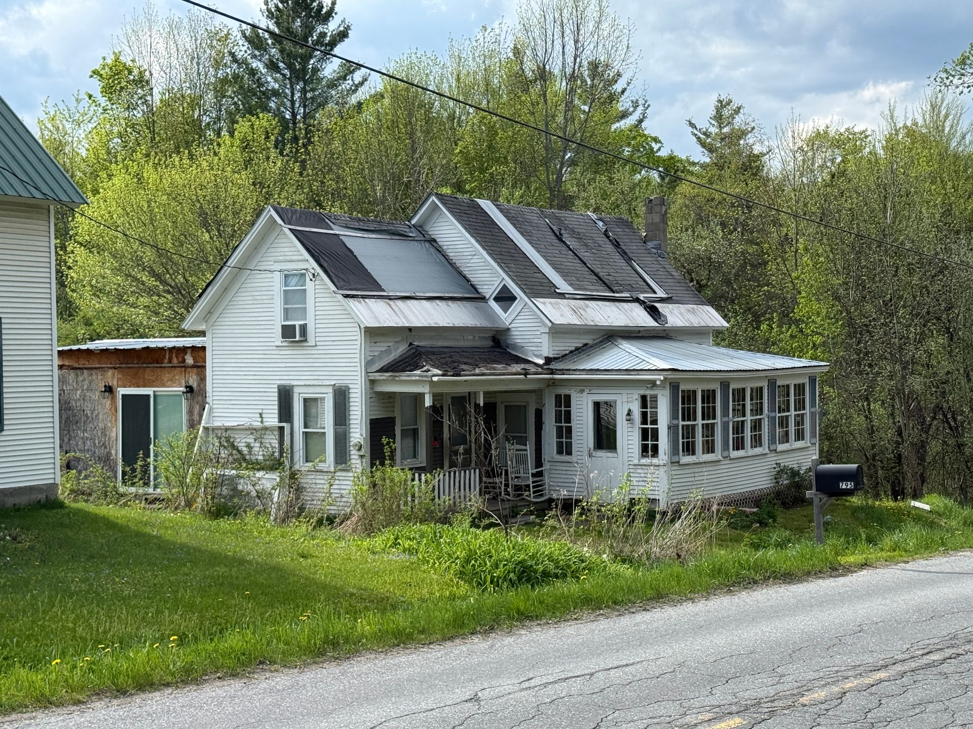 White house with solar panels and porch beside a gravel road, surrounded by trees