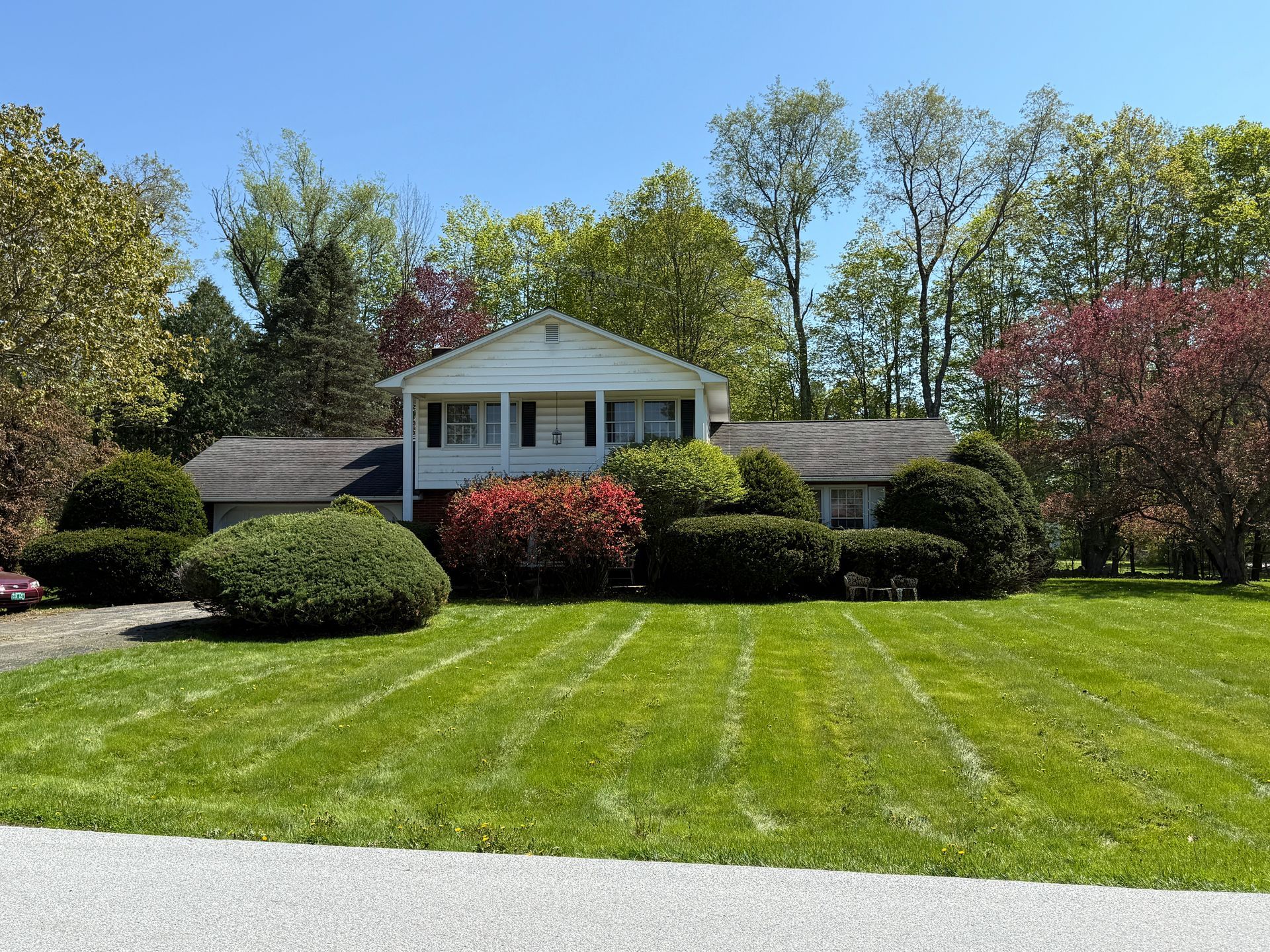 Lawn in front of a white house with a porch, bushes, and trees under a clear blue sky.