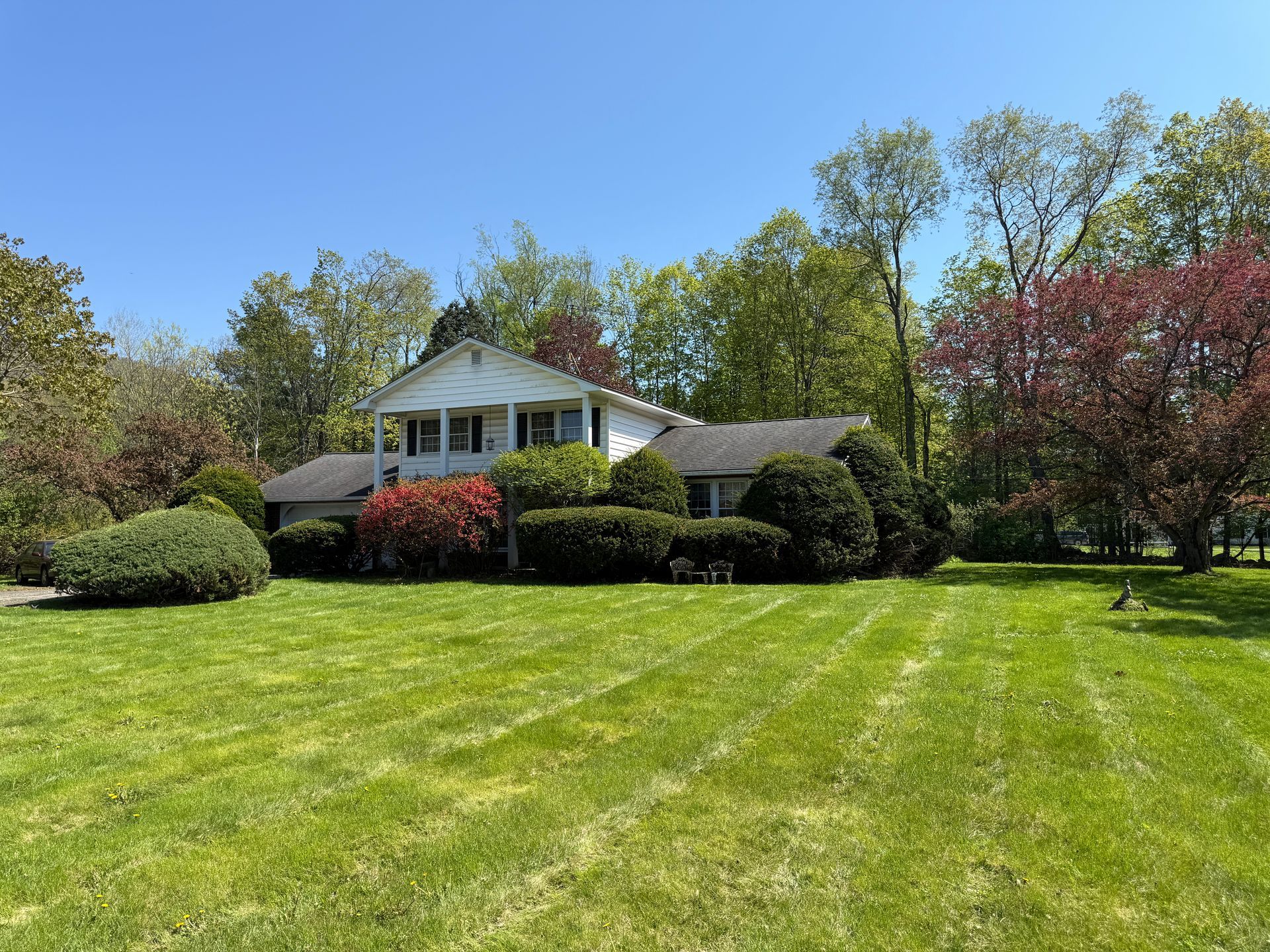White two-story house with green lawn and various colorful shrubs and trees under a clear blue sky.