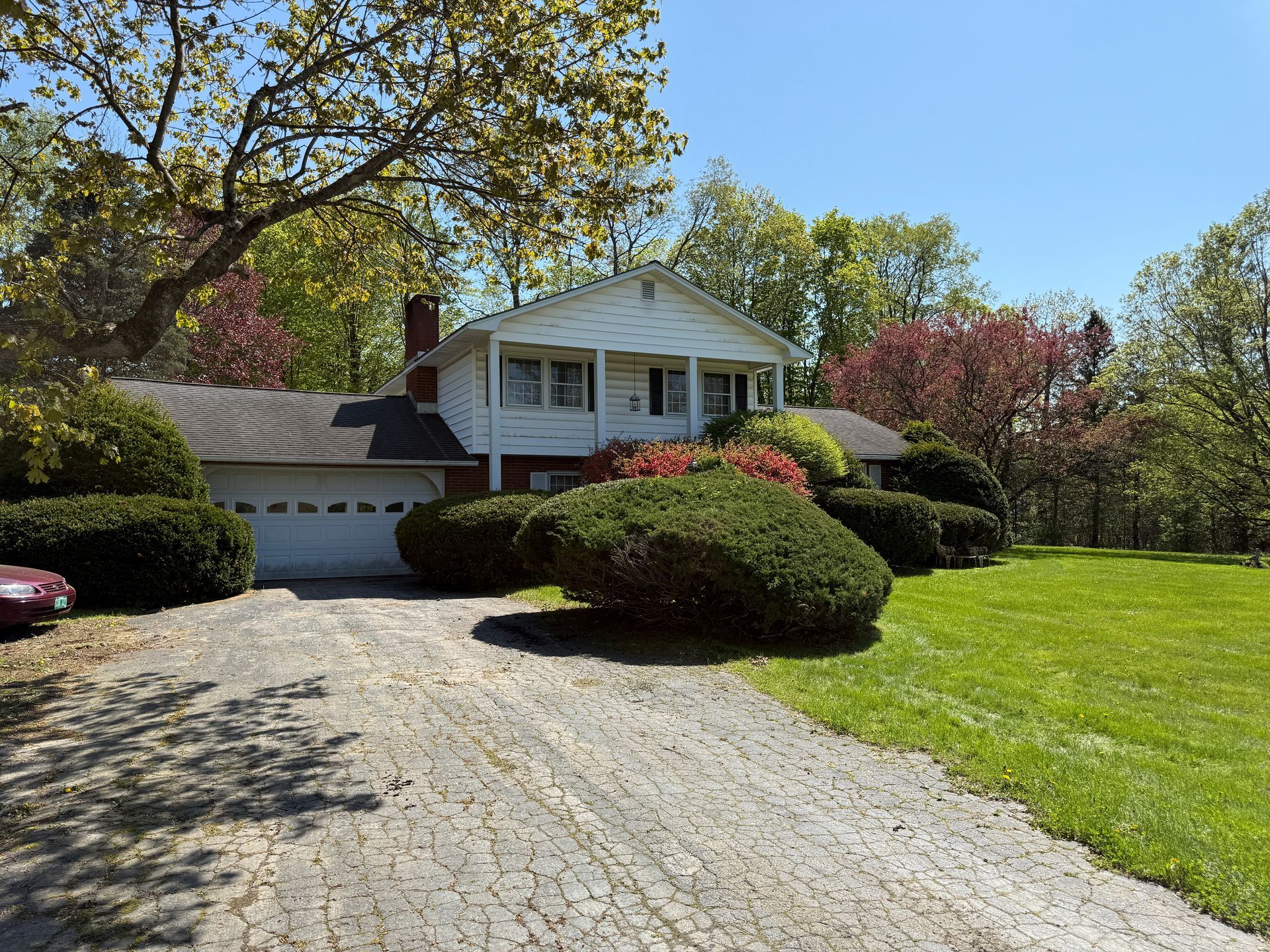 House with white siding and a driveway, surrounded by green shrubs, trees, and grass on a sunny day.