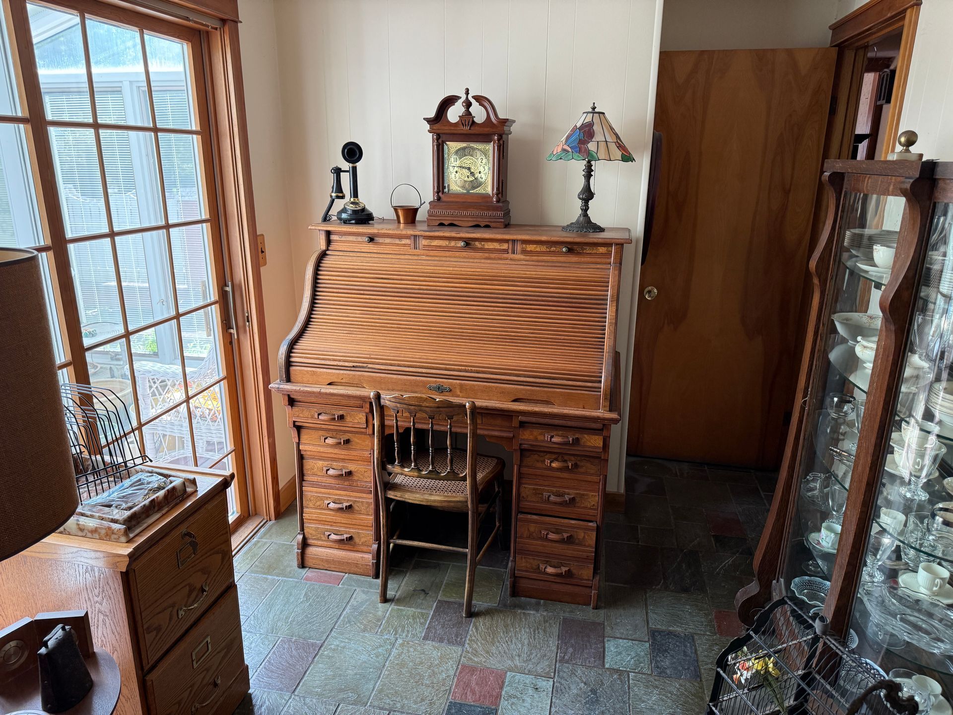 Antique wooden roll-top desk with chair, clock, and display case in a room with a window and a door.