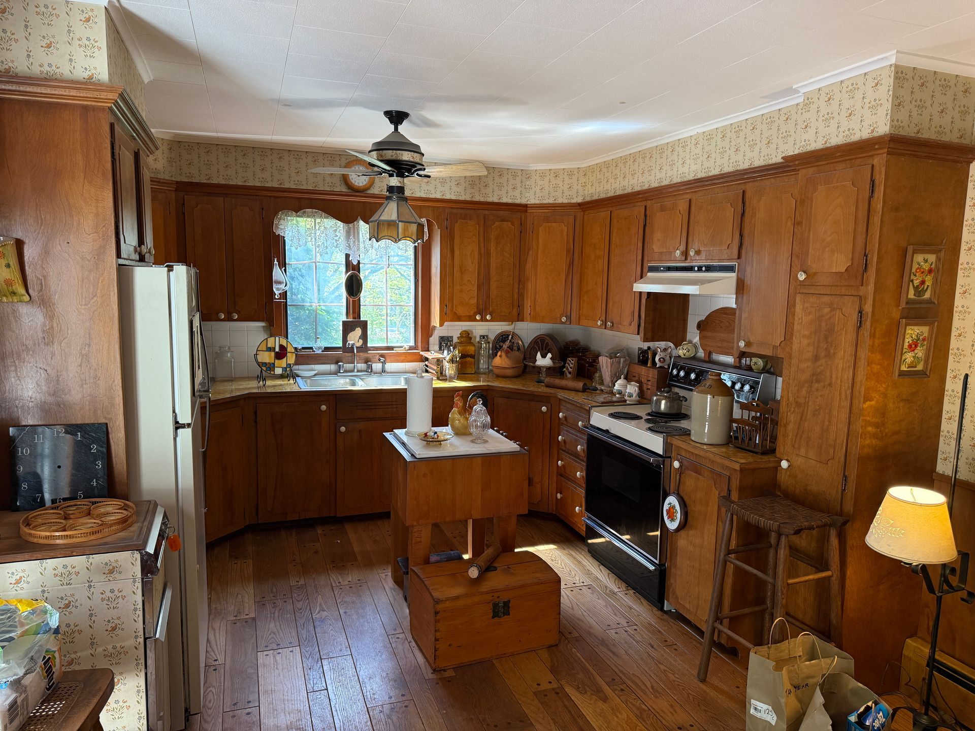 Wooden-cabinet kitchen with a refrigerator, stove, island, window, and ceiling fan. Light wood floors and floral wallpaper.