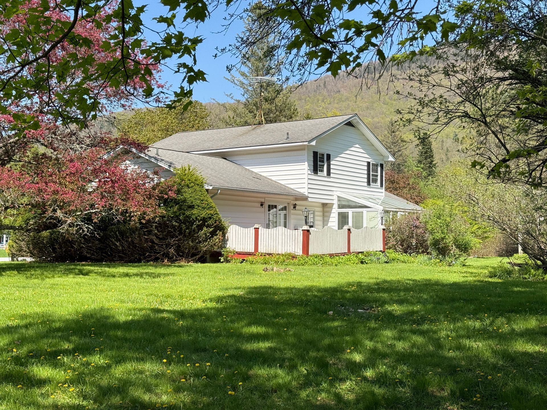 White house with green yard, red trim, trees, and mountain backdrop.
