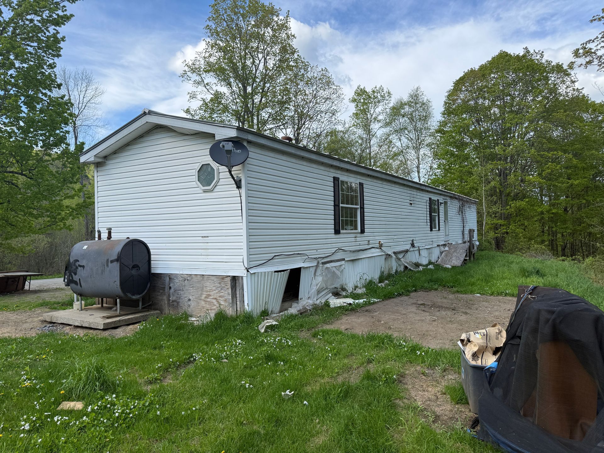 Mobile home, siding, outdoor setting with trees and grass, fuel tank, cloudy sky.