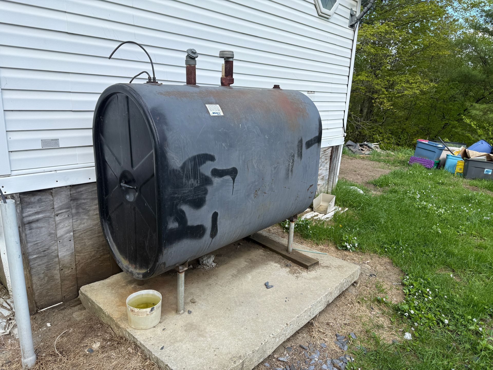 Oil tank on a concrete pad next to a white-sided building. Green grass and blue sky are in the background.