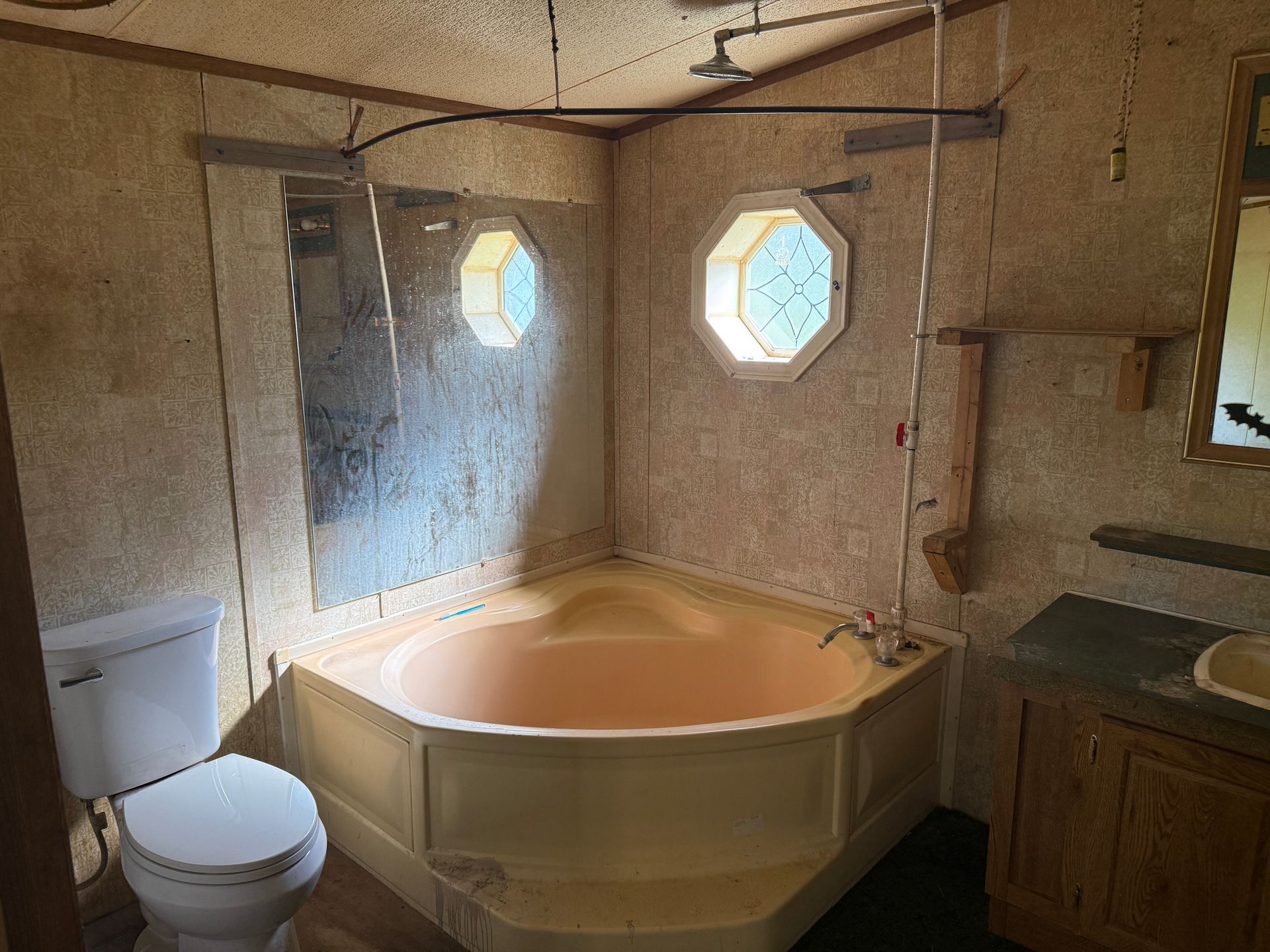 Bathroom with corner tub, toilet, octagonal window, shower head. Light brown walls, wood cabinets.